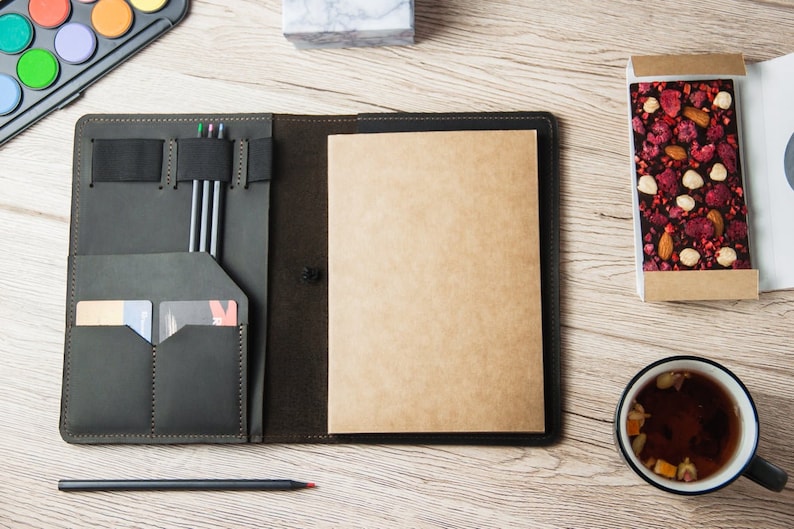 Puede incluir: Un cuaderno de cuero verde oscuro con un portaplumas integrado, ranuras para tarjetas y un cuaderno de papel kraft en blanco. El cuaderno est&aacute; abierto y sobre una mesa de madera con una taza de t&eacute;, una caja de chocolate y un juego de acuarelas.
