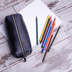 Peut inclure: Une trousse &agrave; crayons en cuir noir avec une fermeture &agrave; glissi&egrave;re, pos&eacute;e sur une surface en bois blanc avec une feuille de papier blanc et une vari&eacute;t&eacute; de crayons de couleur.