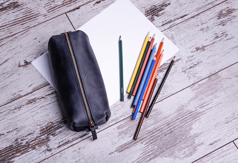 Peut inclure: Une trousse &agrave; crayons en cuir noir avec une fermeture &agrave; glissi&egrave;re, pos&eacute;e sur une surface en bois blanc avec une feuille de papier blanc et une vari&eacute;t&eacute; de crayons de couleur.
