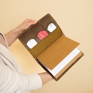 K&ouml;nnte beinhalten: Ein braunes Leder-Notizbuchcover mit integriertem Kartenfach und einem Notizblock mit liniertem Papier.