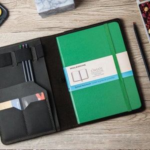 Peut inclure: Une couverture de carnet en cuir noir avec plusieurs poches et fentes pour stylos, cartes et un carnet. La couverture est ouverte pour révéler un carnet Moleskine vert avec le texte "Classic Collection" et "Dotted Notebook" imprimé sur le devant.