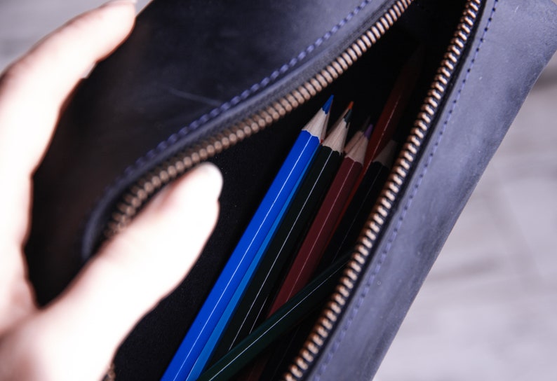 Peut inclure: Une trousse &agrave; crayons en cuir noir avec une fermeture &agrave; glissi&egrave;re. La trousse est ouverte et remplie de crayons de couleur.