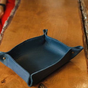 Puede incluir: Una bandeja de cuero azul oscuro con forma cuadrada y esquinas redondeadas. La bandeja est&aacute; hecha de cuero suave y flexible y tiene un brillo sutil. Est&aacute; sobre una superficie de madera.