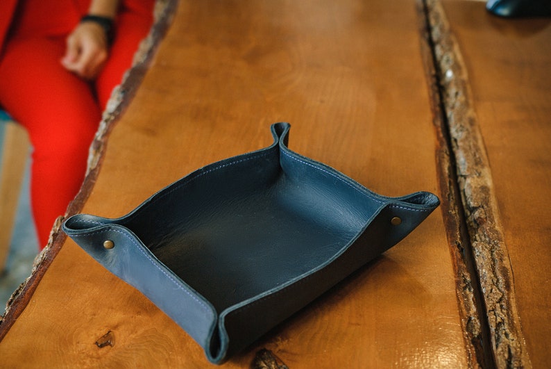 Puede incluir: Una bandeja de cuero azul oscuro con forma cuadrada y esquinas redondeadas. La bandeja est&aacute; hecha de cuero suave y flexible y tiene un brillo sutil. Est&aacute; sobre una superficie de madera.