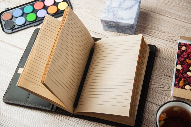 Puede incluir: Un cuaderno abierto con l&iacute;neas, con una cubierta de cuero negro, un juego de pintura de acuarela, una peque&ntilde;a caja de m&aacute;rmol blanco, una barra de chocolate con nueces y frutos secos, y una taza de t&eacute; sobre una mesa de madera.