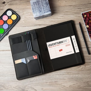 May include: A black leather notebook cover with a built-in pen holder and pockets. The cover is open to reveal a weekly planner from LEUCHTTURM1917, titled "Denken mit der Hand" and dated 2025.