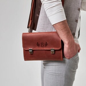 K&ouml;nnte beinhalten: Eine braune Ledertasche mit einem silbernen Verschluss und einem personalisierten Monogramm des Buchstabens "B" mit einem Lorbeerkranz-Design.