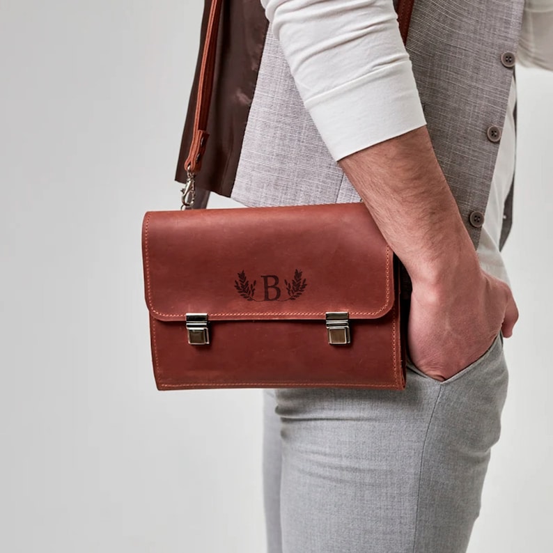 K&ouml;nnte beinhalten: Eine braune Ledertasche mit einem silbernen Verschluss und einem personalisierten Monogramm des Buchstabens "B" mit einem Lorbeerkranz-Design.
