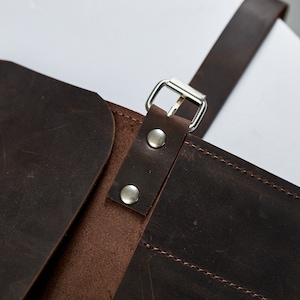 K&ouml;nnte beinhalten: Nahaufnahme einer braunen Ledertasche mit einer silbernen Schnalle und zwei silbernen Nieten. Die Tasche hat eine Klappe, die sich umklappen l&auml;sst und mit der Schnalle befestigt wird.