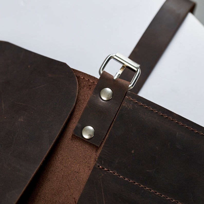 K&ouml;nnte beinhalten: Nahaufnahme einer braunen Ledertasche mit einer silbernen Schnalle und zwei silbernen Nieten. Die Tasche hat eine Klappe, die sich umklappen l&auml;sst und mit der Schnalle befestigt wird.