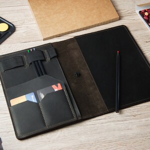 Puede incluir: Una libreta de cuero negro con varios bolsillos y ranuras para bol&iacute;grafos, l&aacute;pices y tarjetas. La libreta est&aacute; abierta y tiene un interior de ante marr&oacute;n.