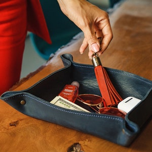 Puede incluir: Una bandeja de cuero azul con un llavero de borla roja, gafas de sol, un tubo de crema de manos y una funda blanca. La bandeja est&aacute; sobre una superficie de madera.