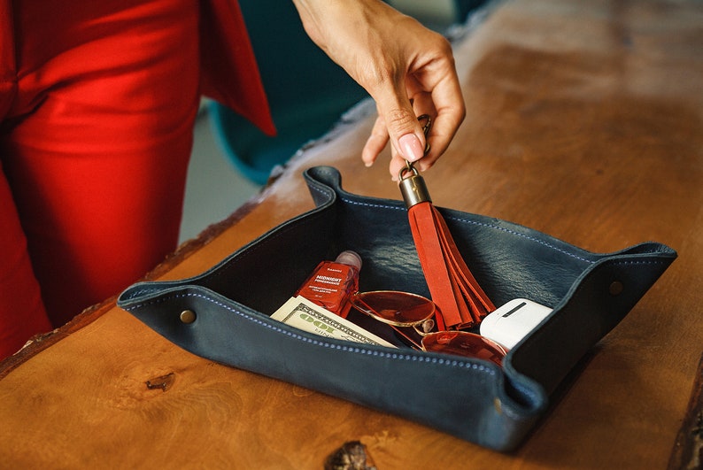 Puede incluir: Una bandeja de cuero azul con un llavero de borla roja, gafas de sol, un tubo de crema de manos y una funda blanca. La bandeja est&aacute; sobre una superficie de madera.