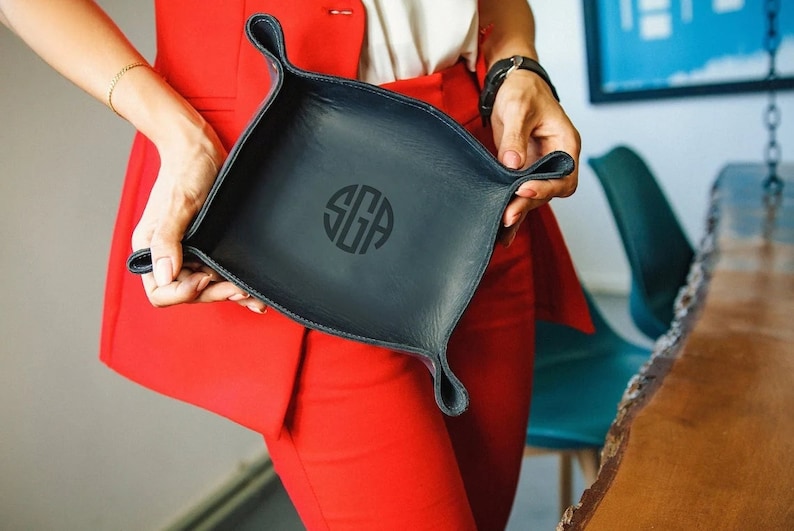 Puede incluir: Una bandeja de cuero negro con un c&iacute;rculo monogramado que dice "SGA".