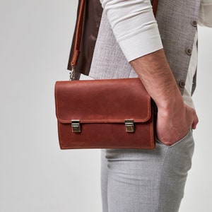 May include: A brown leather briefcase with silver clasps and a strap. The briefcase is being carried by a person wearing a white shirt and gray pants.