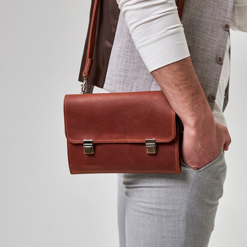 May include: A brown leather briefcase with silver clasps and a strap. The briefcase is being carried by a person wearing a white shirt and gray pants.
