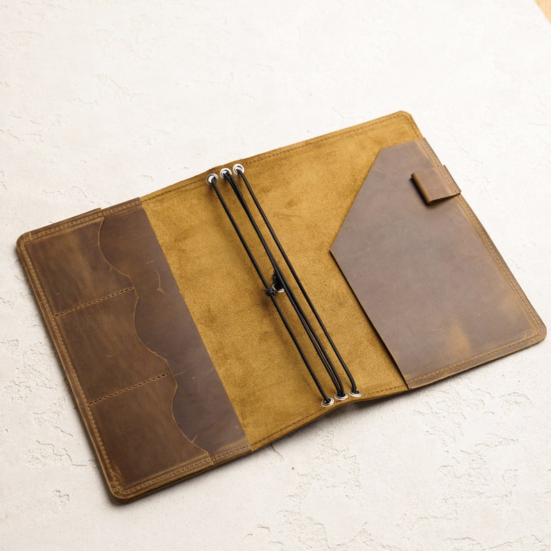 K&ouml;nnte beinhalten: Ein braunes, ledergebundenes Tagebuch mit Klappenverschluss und Gummibandverschluss. Das Tagebuch hat eine Tasche auf der Innenseite des Deckels und ein liniertes Innere.
