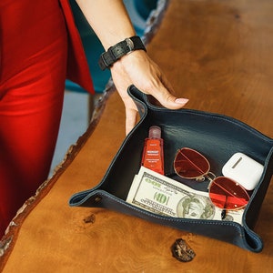 Puede incluir: Una bandeja de cuero azul con varios art&iacute;culos en su interior, incluyendo una peque&ntilde;a botella de desinfectante de manos, unas gafas de sol rojas, una funda blanca y un billete de d&oacute;lar.