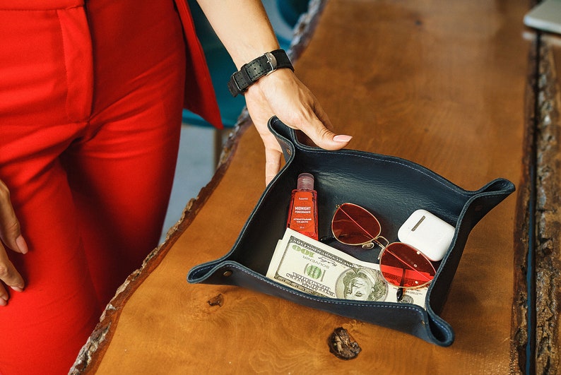 Puede incluir: Una bandeja de cuero azul con varios art&iacute;culos en su interior, incluyendo una peque&ntilde;a botella de desinfectante de manos, unas gafas de sol rojas, una funda blanca y un billete de d&oacute;lar.