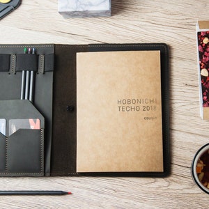 Puede incluir: Una cubierta de agenda de cuero negro con un cuaderno de papel marr&oacute;n en el interior. El cuaderno tiene el texto "HOBONICHI TECHNO 2018 COUSIN" impreso en la portada. La cubierta tiene un portaplumas, ranuras para tarjetas y un cierre de banda el&aacute;stica.