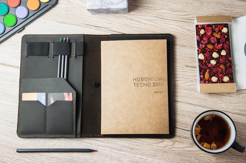 Puede incluir: Una cubierta de agenda de cuero negro con un cuaderno de papel marr&oacute;n en el interior. El cuaderno tiene el texto "HOBONICHI TECHNO 2018 COUSIN" impreso en la portada. La cubierta tiene un portaplumas, ranuras para tarjetas y un cierre de banda el&aacute;stica.