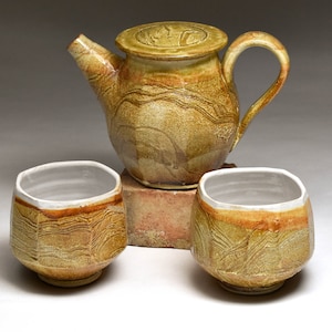 May include: A ceramic teapot and two matching cups in a light brown and yellow glaze with a textured, marbled pattern. The teapot has a lid and a handle. The cups are round and have a white interior.