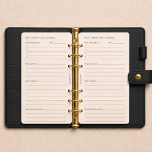 May include: A black leather-textured binder with a gold-toned six-ring metal mechanism holding cream-coloured pages. The pages are printed with Daily Gratitude Journal, Today I'm grateful for, Today's affirmations, Today's reflection, and Tomorrow I look forward to.
