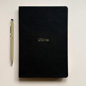 Puede incluir: Un diario negro con el nombre "Olivia" en letras doradas se muestra junto a un bolígrafo dorado. El diario está cerrado y tiene una textura suave. El bolígrafo es delgado y tiene una punta y detalles dorados.