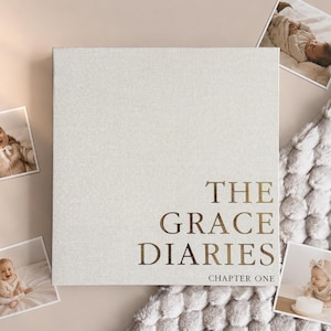 May include: A cream-coloured linen-textured baby memory book with the gold text "THE GRACE DIARIES CHAPTER ONE". The book is surrounded by four small photographs of babies, set against a neutral backdrop.