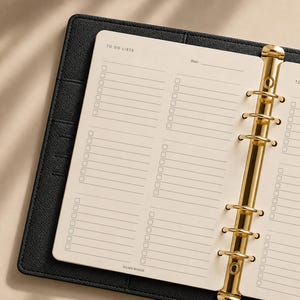 Puede incluir: Una carpeta con textura de cuero negro y un mecanismo metálico dorado de seis anillas que sujeta hojas de papel color crema. Las páginas incluyen el texto impreso TO DO LISTS y Date, con varias columnas de líneas y casillas de verificación.