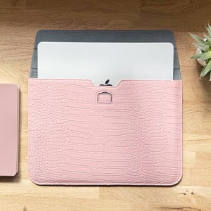 Op de afbeelding: Een roze laptophoes met een krokodillenleertextuur. De hoes is open en toont een witte laptop met een zwart appel-logo. De binnenkant is grijs. Links is een klein, bijpassend roze item zichtbaar.