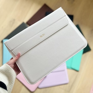 Op de afbeelding: Een lichtbeige laptophoes met een klepsluiting. De initialen "SLJU" zijn in goud op de voorkant geëmbosseerd. Andere gekleurde hoezen zijn zichtbaar op de achtergrond, waaronder roze, groen en bruin.