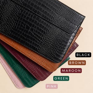 Op de afbeelding: Een stapel laptophoezen van imitatieleer in verschillende kleuren, waaronder zwart, bruin, bordeauxrood, groen en roze. Elke hoes heeft een textuur met krokodillenpatroon. De kleurnamen worden verticaal weergegeven.