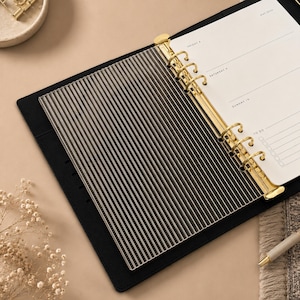 Könnte beinhalten: Schwarzer Lederplaner mit goldfarbenen Metallringen und einem gestreiften Trennblatt. Der Planer ist auf einer Seite mit den Wochentagen und einer To-Do-Liste geöffnet. Der Hintergrund ist ein neutrales Beige.
