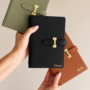 May include: Three leather passport holders in olive green, black, and brown. Each has a gold-coloured buckle closure and personalised text. The black passport holder is labelled "Grace", and the brown one is labelled "SLJU".