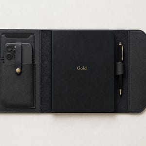 May include: A black folio organiser featuring a central notebook with the word Gold embossed in gold lettering. The left side includes a smartphone pocket with a snap closure, while the right side holds a black pen in a dedicated loop.