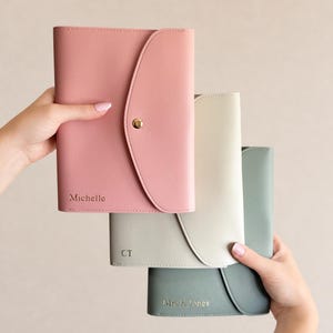 Op de afbeelding: Drie rechthoekige planners van kunstleer met afgeronde klepsluitingen en goudkleurige drukknopen. De bovenste planner is roze met de naam Michelle, de middelste is crème met de initialen CT en de onderste is saliegroen met Mrs. K Jones.
