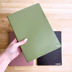 K&ouml;nnte beinhalten: Eine Hand h&auml;lt ein gr&uuml;nes Notizbuch, mit einem rosa und schwarzen Notizbuch im Hintergrund. Auf dem schwarzen Notizbuch steht der Name "Andrew". Das gr&uuml;ne Notizbuch hat die Initialen "BK". Das rosa Notizbuch hat den Namen "Morgan".