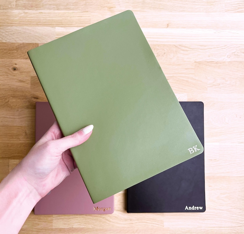 K&ouml;nnte beinhalten: Eine Hand h&auml;lt ein gr&uuml;nes Notizbuch, mit einem rosa und schwarzen Notizbuch im Hintergrund. Auf dem schwarzen Notizbuch steht der Name "Andrew". Das gr&uuml;ne Notizbuch hat die Initialen "BK". Das rosa Notizbuch hat den Namen "Morgan".