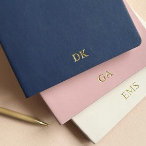 Op de afbeelding: Drie gepersonaliseerde notitieboekjes in marineblauw, roze en wit, elk met gouden initialen. Een gouden pen ligt ernaast. De notitieboekjes zijn een stijlvol briefpapierartikel.