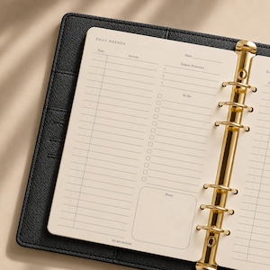 Può includere: Un raccoglitore in pelle nera con un meccanismo a sei anelli color oro che trattiene pagine di agenda giornaliera color crema. Le pagine presentano sezioni per ora, attività, data, priorità, una lista di cose da fare con caselle e un riquadro per le note, con HUMEWOOD stampato in basso.