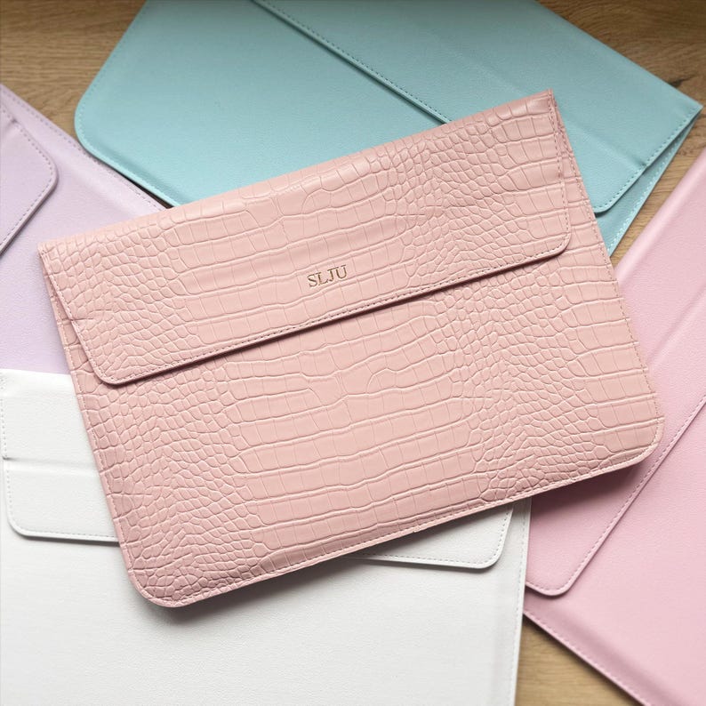 Op de afbeelding: Een roze laptoptas met een krokodillenleertextuur en een klepsluiting. De initialen "SLJU" zijn op de voorkant in reli&euml;f aangebracht. Andere pastelkleurige laptoptassen zijn op de achtergrond zichtbaar.