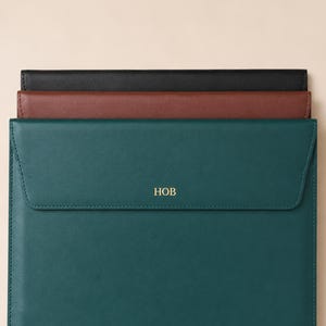Peut inclure: Trois pochettes pour ordinateur portable en cuir noir, marron et vert d'eau. La pochette verte d'eau a une fermeture à rabat et les lettres dorées "HOB" en relief sur le devant. Les pochettes sont empilées les unes sur les autres.