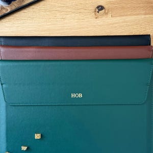 以下が含まれることがあります： 黒、茶色、ティールグリーンの3色の革製ラップトップスリーブが木製の表面に重ねられています。ティールグリーンのスリーブには金色のイニシャル「HOB」と小さな金色の装飾が施されています。スリーブはフラップで閉じられています。
