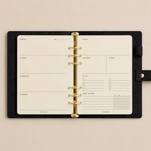 Peut inclure: Un agenda à reliure en cuir noir avec un mécanisme de reliure métallique doré à six anneaux. Les pages ouvertes présentent du papier crème avec des sections imprimées pour lundi à dimanche, une liste de tâches et un suivi d'habitudes.