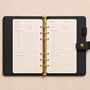 Op de afbeelding: Een zwarte map met ledertextuur en een goudkleurig mechanisme met zes ringen, waarin twee crèmekleurige receptpagina's zitten. Elke pagina bevat gedrukte velden voor recepttitel, datum, voorbereidingstijd, kooktijd, porties, ingrediënten en instructies.