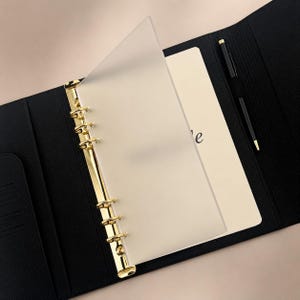 Op de afbeelding: Zwarte leren ordner met goudkleurige metalen ringen en accenten. De ordner is open en toont crèmekleurig papier en een transparante plastic scheidingswand. Twee zwarte pennen zijn zichtbaar.