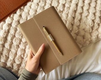 Peut inclure: Un carnet en cuir beige avec un stylo doré assorti. Le carnet est fermé et tenu dans une main. Le stylo repose sur le carnet. Le fond est un plaid blanc et moelleux.