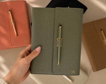 May include: A collection of rectangular notebooks in various colours including olive green, coral, tan, black, and cream. Each notebook features a textured cover, a gold-toned pen holder with a matching pen, and gold-embossed initials on the front.