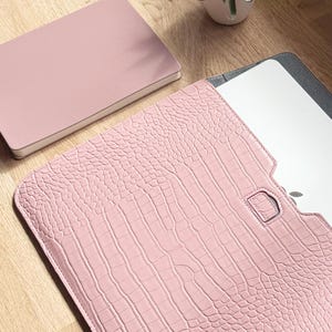 Op de afbeelding: Een roze laptoptas met een krokodillenleertextuur is open en onthult een laptop. Een bijpassend roze notitieboekje ligt naast de hoes. De items liggen op een houten oppervlak. De hoes heeft een D-ringsluiting.